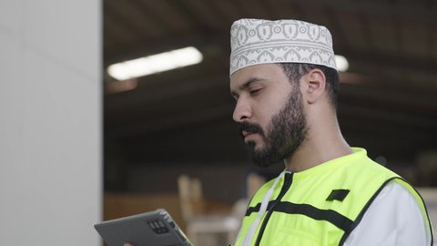 التحول التقني والتكنولوجي الحديث, اعتماد الآليات و الذكاء الاصطناعي في الهندسة, تشغيل خط الإنتاج بإستخدام جهاز التابلت, تصوير مقرب لمهندس عربي  خليجي عماني يرتدي السترة الحماية و الكمة يتابع سير الأعمال داخل المصنع