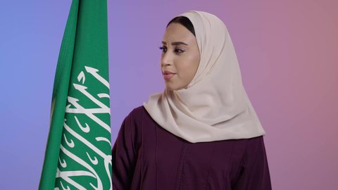 النظر الى الكاميرا بايماءات الفخر لامتلاك الهوية السعودية, تخليد يوم العلم 11 مارس, الاحتفال بذكرى اليوم الوطني السعودي 23 سبتمبر, بورترية لامرأة عربية سعودية خليجية مبتمسة ترتدي العباءة والحجاب تمسك علم المملكة ، خلفية ملونه