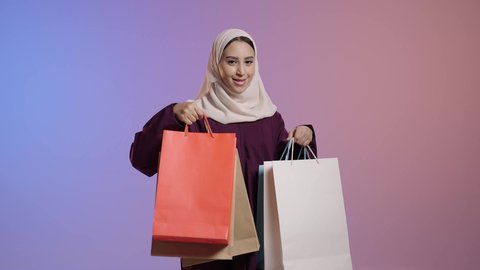 ايماءات السعاده و السرور، التسوق من المولات التجارية، استغلال العروض والتخفيضات الموسمية والجمعة البيضاء، الترويج لماركات عالمية فاخرة، بورترية لإمرأة عربية خليجية سعودية محجبة ترتدي العباءه تحمل بيدها أكياس ورقية، خلفية ملونه