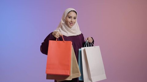 التسوق من المولات التجارية، استغلال العروض والتخفيضات الموسمية والجمعة البيضاء، الترويج لماركات عالمية فاخرة، بورترية لإمرأة عربية خليجية سعودية محجبة ترتدي العباءه تحمل بيدها أكياس ورقية، ايماءات السعاده و السرور، خلفية ملونه
