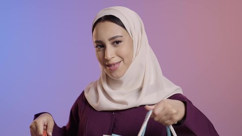استغلال العروض والتخفيضات الموسمية والجمعة البيضاء، الترويج لماركات عالمية فاخرة، بورترية مقرب لإمرأة عربية خليجية سعودية محجبة ترتدي العباءه تحمل بيدها أكياس ورقية، التسوق من المولات التجارية، ايماءات السعاده و السرور، خلفية ملونه