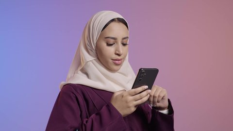 الدردشة مع الأصدقاء والأقارب عبر الجوال، تصفح مواقع التواصل الاجتماعي، استخدام جهاز تقني حديث ومتطور، بورتريه لشابة عربيه خليجيه سعودية ترتدي الحجاب و العباءة تمسك بيدها الهاتف المحمول، خلفية ملونه