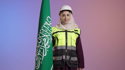 الاحتفال بذكرى اليوم الوطني السعودي 23 سبتمبر، تخليد يوم العلم 11 مارس، النظر الى الكاميرا بايماءات الفخر لامتلاك الهوية السعودية, بورترية لمهندسة عربية سعودية خليجية مبتمسة ترتدي خوذه و ستره الحماية تمسك علم المملكة ، خلفية ملونه