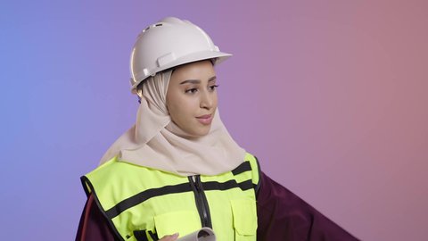 دور الهندسة الإنشائية في تطور المجتمع السعودي، حمل المخطط الهندسي،, بورتريه مقرب لمهندسة عربية خليجية سعودية مسلمة محجبة ترتدي الخوذة البيضاء وسترة الحماية تشير الى شيء ما، خلفية ملونه