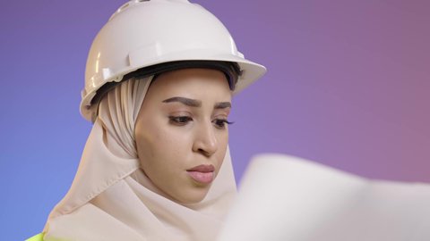 مفهوم الهندسة وأعمال البناء, بورتريه مقرب لمهندسة عربية خليجية سعودية محجبة ترتدي عباءة سوداء مع السترة وخوذة الحماية تقوم بدراسه وتصفح المخططات الهندسيه، مهن ووظائف نسائية، العمل في القطاع الهندسي في السعودية, خلفية ملونه