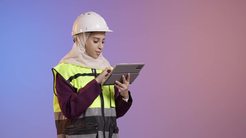 تدوين و تسجيل الملاحظات، الأجهزة التقنية الحديثة، التأكد من سير الأعمال بالشكل المطلوب والصحيح، كتابة آخر المستجدات حول المشروع، بورتريه لمهندسة عربية خليجية سعودية ترتدي الحجاب والخوذة وسترة الحماية تقوم باستخدام الجهاز اللوحي, خلفية ملونه
