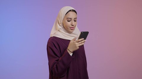 محادثات أونلاين مع الأصدقاء، متابعة أخبار وسائل التواصل الاجتماعي باستخدام الجوال، بورتريه لإمرأة عربية خليجية سعودية ترتدي حجاب وعباءة  تمسك بيديها الهاتف المحمول، خلفية ملونه