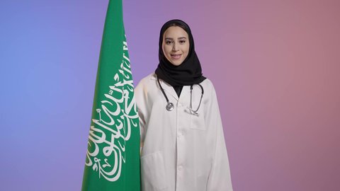Feelings of loyalty to the homeland, Flag Day on March 11, the celebration of the Saudi National Day on September 23, looking at the camera with gestures of pride for possessing the Saudi identity, a portrait of a smiling Arab Saudi Gulf doctor wearing a white coat and stethoscope holding the Kingdom's flag, with a colorful background.