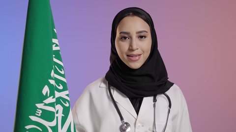 الاعتزاز بالوطن والانتماء له، يوم العلم 11 مارس، الاحتفال بذكرى اليوم الوطني السعودي 23 سبتمبر، النظر الى الكاميرا بايماءات الفخر لامتلاك الهوية السعودية, بورترية مقرب لطبيبة عربية سعودية خليجية مبتمسة ترتدي المعطف الابيض و السماعات الطبية تمسك علم المملكة ، خلفية ملونه