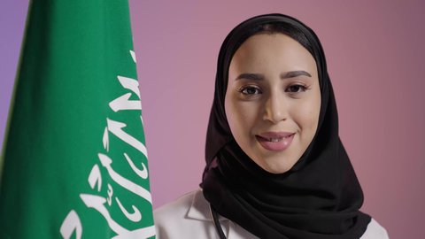 حب الوطن والانتماء إليه، يوم العلم 11 مارس، الاحتفال بذكرى اليوم الوطني السعودي 23 سبتمبر، النظر الى الكاميرا بايماءات الفخر لامتلاك الهوية السعودية, بورترية مقرب لطبيبة عربية سعودية خليجية مبتمسة ترتدي المعطف الابيض و السماعات الطبية تمسك علم المملكة ، خلفية ملونه