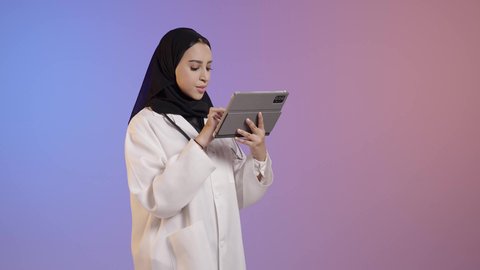 دمج العمل مع التقنية و التكنولوجي، مفهوم الطب و الرعاية الصحية، بورترية لطبيبة عربية خليجية سعودية محجبة ترتدي المعطف الطبي والسماعات الطبية تحمل بيدها جهاز التابلت، خلفية ملونه