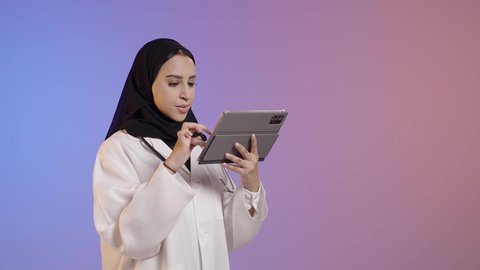 The concept of medicine and healthcare, a portrait of a veiled Arab Gulf Saudi female doctor wearing a medical coat and stethoscope, holding a tablet in her hand, integrating work with technology, colorful background.
