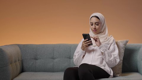 تبادل الأحاديث والرسائل عبر الجوال، استخدام جهاز تقني حديث ومتطور، بورتريه لشابة عربيه خليجيه سعودية ترتدي الحجاب و العباءة تجلس على الاريكة تمسك بيدها الهاتف المحمول، خلفية ملونه