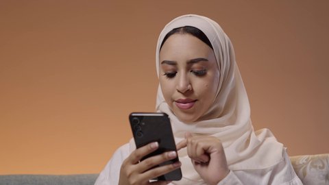 استخدام الجوال للتواصل مع الأصدقاء والأهل، استخدام جهاز تقني حديث ومتطور، بورتريه مقرب لشابة عربيه خليجيه سعودية ترتدي الحجاب و العباءة تمسك بيدها الهاتف المحمول، خلفية ملونه