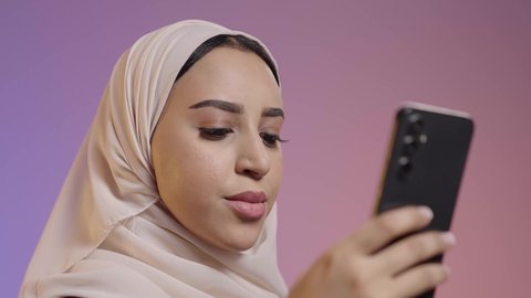 تبادل الرسائل والدردشة عبرالجوال، تصفح مواقع التواصل الاجتماعي، استخدام جهاز تقني حديث ومتطور، بورتريه مقرب لشابة عربيه خليجيه سعودية ترتدي الحجاب و العباءة تمسك بيدها الهاتف المحمول، خلفية ملونه