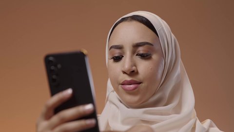 محادثات مع الأقارب والأصدقاء باستخدام الجوال، تصفح مواقع التواصل الاجتماعي، استخدام جهاز تقني حديث ومتطور، بورتريه مقرب لشابة عربيه خليجيه سعودية ترتدي الحجاب و العباءة تمسك بيدها الهاتف المحمول، خلفية ملونه
