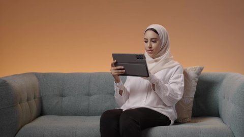 The concept of remote project management, expressions and gestures of integration, the use of advanced modern technology in professional life via the tablet, a portrait of a young Arab Gulf Saudi woman wearing a hijab and abaya sitting on the sofa working on a tablet, colorful background.