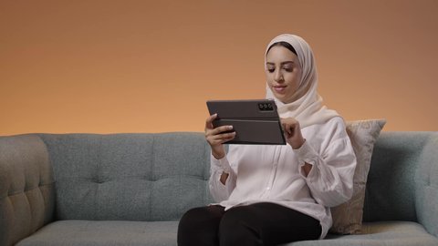 Browsing social media, expressions and gestures of integration, using advanced modern technology in professional life via a tablet, a portrait of a young Arab Gulf Saudi woman wearing a hijab and abaya sitting on a sofa working on a tablet, colorful background.