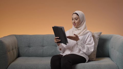 اجراء مكالمه فيديو عبر الجهاز اللوحي، الدردشة مع الأهل والأصدقاء وتصفح مواقع التواصل الاجتماعي، مشاهدة الأفلام والمسلسلات في أوقات الفراغ، بورتريه لامرأة عربية خليجية سعودية محجبه ترتدي العباءة تجلس على الاريكة تمسك الجهاز اللوحي، استخدام  الأجهزة التقنية الحديثة، خلفية ملونه