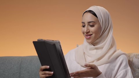Chatting with family and friends and browsing social media, making a video call via the tablet, watching movies and series in free time, a close-up portrait of a Saudi Arabian Gulf woman wearing a hijab and an abaya sitting on the sofa holding a tablet, using modern technological devices, colorful background.