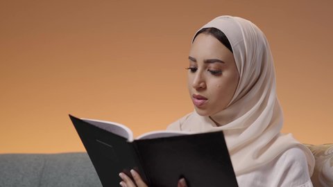 هواية القراءة والاستكشاف، قضاء وقت الفراغ بالتعلم والثقافة، مفهوم التثقيف والبحث العلمي، بورترية مقرب لشابة عربية خليجية سعودية ترتدي العباية والحجاب تجلس على كنبة مريحة تقوم بقراءة كتاب ما، خلفية ملونه