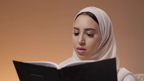 قضاء وقت الفراغ بالتعلم والثقافة، هواية القراءة والاستكشاف، مفهوم التثقيف والبحث العلمي، بورترية لشابة عربية خليجية سعودية ترتدي العباية والحجاب تجلس على كنبة مريحة تقوم بقراءة كتاب ما، خلفية ملونه