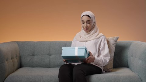 هدية للأعياد والمناسبات السعيدة، إيماءات السعادة والدهشة بالحصول على هدية ثمينة، مفهوم المفاجأة والإهداء، بورتريه لامراة عربيه خليجيه سعودية محجبه ترتدي العباءة تجلس على الاريكة تقوم بمشاهده العلبة بتعابير الفرح والسرور، خلفية ملونه