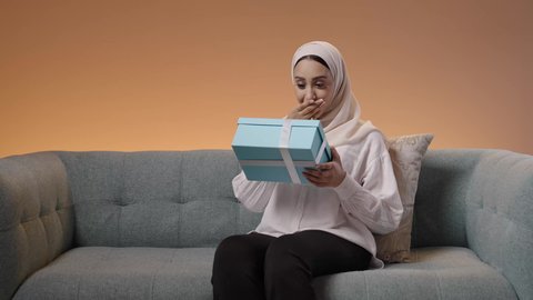 فن انتقاء الهدايا بعناية، إيماءات السعادة والدهشة بالحصول على هدية ثمينة، مفهوم المفاجأة والإهداء، بورتريه لامراة عربيه خليجيه سعودية محجبه ترتدي العباءة تجلس على الاريكة تقوم بمشاهده العلبة بتعابير الفرح والسرور، خلفية ملونه