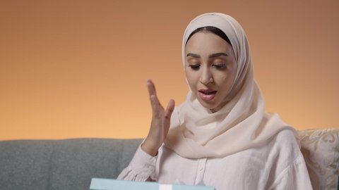 Gestures of happiness and surprise at receiving a precious gift, the concept of surprise and gifting, the art of carefully selecting gifts, a close-up portrait of a Saudi Arabian Gulf woman wearing a hijab and an abaya sitting on a sofa, looking at the box with expressions of joy and delight, colorful background.