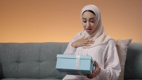 The concept of surprise and gifting, gestures of happiness and astonishment at receiving a precious gift, the art of carefully selecting gifts, a portrait of a Saudi Arabian Gulf woman wearing a hijab and an abaya sitting on a sofa, watching the box with expressions of joy and delight, colorful background.