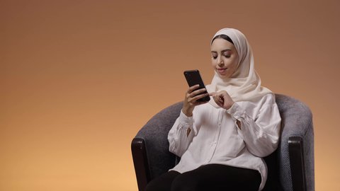 التواصل الإلكتروني مع الأصدقاء، متابعة أخبار وسائل التواصل الاجتماعي باستخدام الجوال، بورتريه لإمرأة عربية خليجية سعودية ترتدي حجاب وعباءة تجلس على كرسي تمسك بيديها الهاتف المحمول، خلفية ملونه
