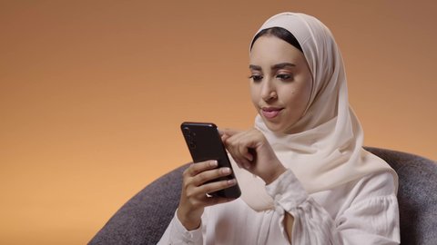 متابعة أخبار وسائل التواصل الاجتماعي باستخدام الجوال، بورتريه مقرب لإمرأة عربية خليجية سعودية ترتدي حجاب وعباءة تجلس على كرسي تمسك بيديها الهاتف المحمول، الدردشة والتواصل مع الأصدقاء أونلاين، خلفية ملونه