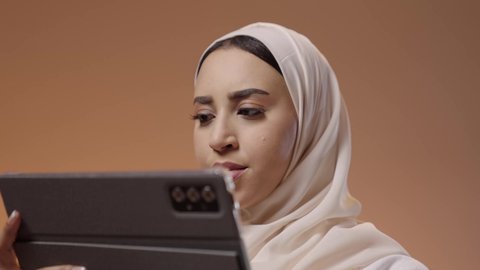 Touching the screen to navigate between applications, browsing social media, expressions and gestures of integration, using advanced modern technology in professional life, a close-up portrait of a young Arab Gulf Saudi woman wearing a hijab and abaya working on a tablet, colorful background.