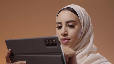 استخدام شاشة الجهاز اللوحي بالنقر والتحريك، تصفح مواقع التواصل الاجتماعي، تعابير وايماءات الاندماج، استخدام التقنية الحديثه المتطورة في الحياة العملية، بورتريه مقرب لشابة عربية خليجية سعودية ترتدي الحجاب والعباءة تقوم بالعمل على جهازالتابلت، خلفية ملونه