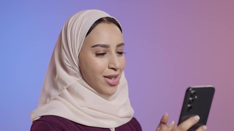 Chatting with friends and relatives via mobile, browsing social media, using a modern and advanced technical device, a close-up portrait of a young Arab Gulf Saudi woman wearing a hijab and abaya holding a mobile phone in her hand, colorful background.