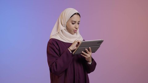 استخدام التقنية الحديثه المتطورة في الحياة العملية عبر الجهاز اللوحي، تعابير وايماءات الاندماج، بورتريه لشابة عربية خليجية سعودية ترتدي الحجاب والعباءة تقوم بالعمل على جهازالتابلت، خلفية ملونه