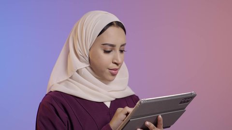 تعابير وايماءات الاندماج، استخدام التقنية الحديثه المتطورة في الحياة العملية عبر الجهاز اللوحي، بورتريه مقرب لشابة عربية خليجية سعودية ترتدي الحجاب والعباءة تقوم بالعمل على جهازالتابلت، خلفية ملونه