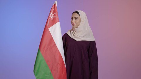 حب الوطن والانتماء إليه، ثقافات و رموز وطنية، العلم الوطني للبلاد، تقديم الولاء للراية العمانية بمشاعر العز والكرامة، بورترية لامراة عربية خليجية عمانية مبتسمة محجبة ترتدي الحجاب و العباءة  تنظر الى الكاميرا بايماءات الفخر، إحياء ذكرى تأسيس الدولة 1775 م، خلفية ملونه