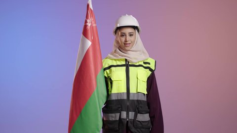 إحياء ذكرى تأسيس الدولة 1775 م، الفخر والاعتزاز بالانتماء لسلطنة عمان، تقديم الولاء للراية العمانية بمشاعر العز والكرامة، بورترية لمهندسة عربية خليجية سعودية مبتسمة محجبة ترتدي سترة و خوذة الحماية تمسك علم عمان، العلم الوطني للبلاد، خلفية ملونه