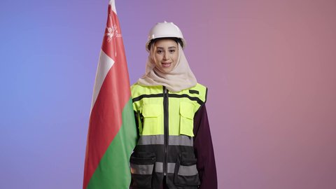 الفخر والاعتزاز بالانتماء لسلطنة عمان، تقديم الولاء للراية العمانية بمشاعر العز والكرامة، بورترية لمهندسة عربية خليجية سعودية مبتسمة محجبة ترتدي سترة و خوذة الحماية تمسك علم عمان، إحياء ذكرى تأسيس الدولة 1775 م، العلم الوطني للبلاد، خلفية ملونه