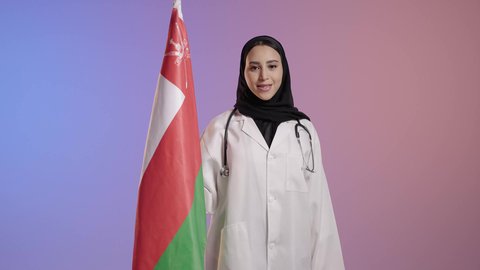 تقديم الولاء للراية العمانية بمشاعر العز والكرامة، الفخر والاعتزاز بالانتماء لسلطنة عمان، بورترية لطبيبة عربية خليجية سعودية مبتسمة محجبة ترتدي المعطف الطبي تمسك علم عمان، إحياء ذكرى تأسيس الدولة 1775 م، العلم الوطني للبلاد، خلفية ملونه