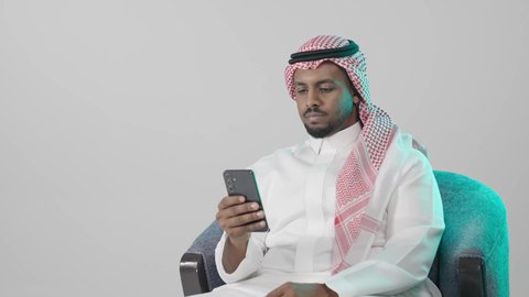 متابعة الأحداث اليومية عبر الشبكات الاجتماعية، بورترية مقرب لرجل عربي خليجي سعودي يرتدي الثوب التقليدي و الشماغ يجلس على كرسي يمسك بيده الهاتف المحمول، الدردشة والتواصل مع الأهل و الأصدقاء، استخدام جهاز تقني حديث ومتطور، العمل عن بعد وانجاز المهام المطلوبة، خلفية رمادية