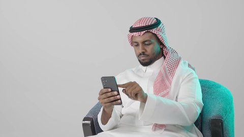 متابعة الأصدقاء أونلاين، بورترية مقرب لرجل عربي خليجي سعودي يرتدي الثوب التقليدي و الشماغ يجلس على كرسي يمسك بيده الهاتف المحمول، الدردشة والتواصل مع الأهل و الأصدقاء، استخدام جهاز تقني حديث ومتطور، العمل عن بعد وانجاز المهام المطلوبة، خلفية رمادية