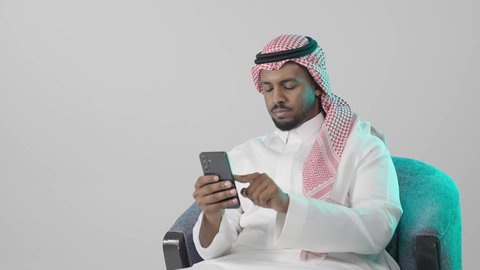 Browse the content of various social media platforms, a close-up portrait of a Saudi Gulf Arab man wearing traditional attire and a ghutrah sitting on a chair holding a mobile phone, chatting and communicating with family and friends, using a modern and advanced technological device, working remotely and completing required tasks, with a gray background.