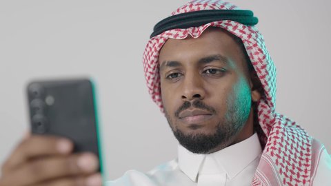 استخدام تطبيقات الشبكات الاجتماعية، بورترية مقرب لرجل عربي خليجي سعودي يرتدي الثوب التقليدي و الشماغ  يمسك بيده الهاتف المحمول، الدردشة والتواصل مع الأهل و الأصدقاء، استخدام جهاز تقني حديث ومتطور، العمل عن بعد وانجاز المهام المطلوبة، خلفية رمادية