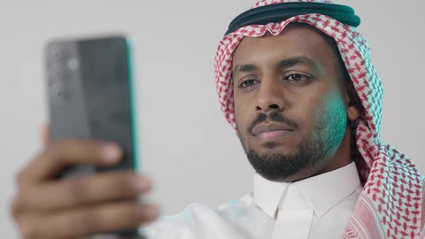 Navigating through social media pages, a close-up portrait of a Saudi Arabian Gulf man wearing traditional attire and a ghutrah, holding a mobile phone in his hand, chatting and communicating with family and friends, using a modern and advanced technological device, working remotely and completing required tasks, gray background.