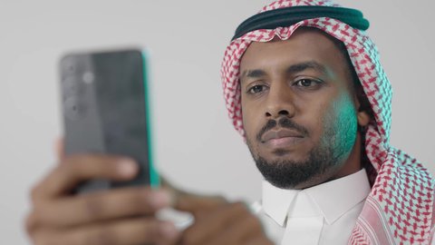 استعراض المنشورات والتحديثات على الإنترنت، بورترية مقرب لرجل عربي خليجي سعودي يرتدي الثوب التقليدي و الشماغ  يمسك بيده الهاتف المحمول، الدردشة والتواصل مع الأهل و الأصدقاء، استخدام جهاز تقني حديث ومتطور، العمل عن بعد وانجاز المهام المطلوبة، خلفية رمادية