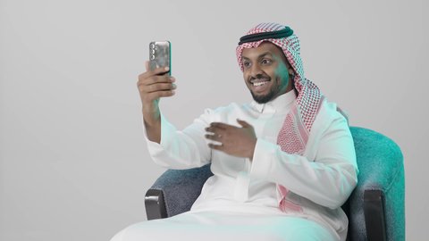 Interacting with family and friends online, hand gestures indicating welcome, making a video call via mobile, a portrait of a Saudi Arabian Gulf man wearing traditional attire and a ghutrah sitting on a chair holding a mobile phone, expressions of happiness and joy, gray background.