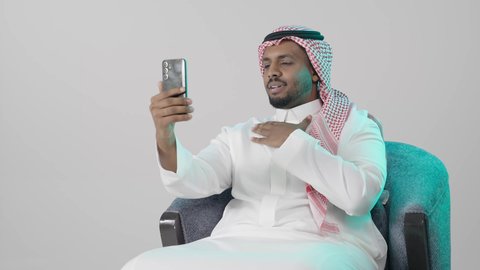 الدردشة مع الأهل والأصدقاء، عرض بث مباشر على وسائل السوشل ميديا، ايماءات يدين تدل على الترحيب، إجراء مكالمة فيديو عبر الجوال , بورتريه لرجل عربي خليجي سعودي  يرتدي الثوب التقليدي و الشماغ يجلس على كرسي يمسك بيده الهاتف المحمول ,تعابيرالسعادة و السرور ، خلفية رمادية