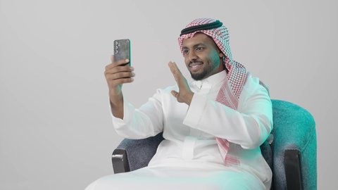 Live broadcast on social media, hand gestures indicating welcome, connecting with family and friends during leisure time, making a video call via mobile, a portrait of a Saudi Arabian Gulf man wearing traditional attire and a ghutrah sitting on a chair holding a mobile phone, expressions of happiness and joy, gray background.